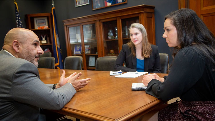 Image of three people sitting at a wood table talking with papers spread out between them.